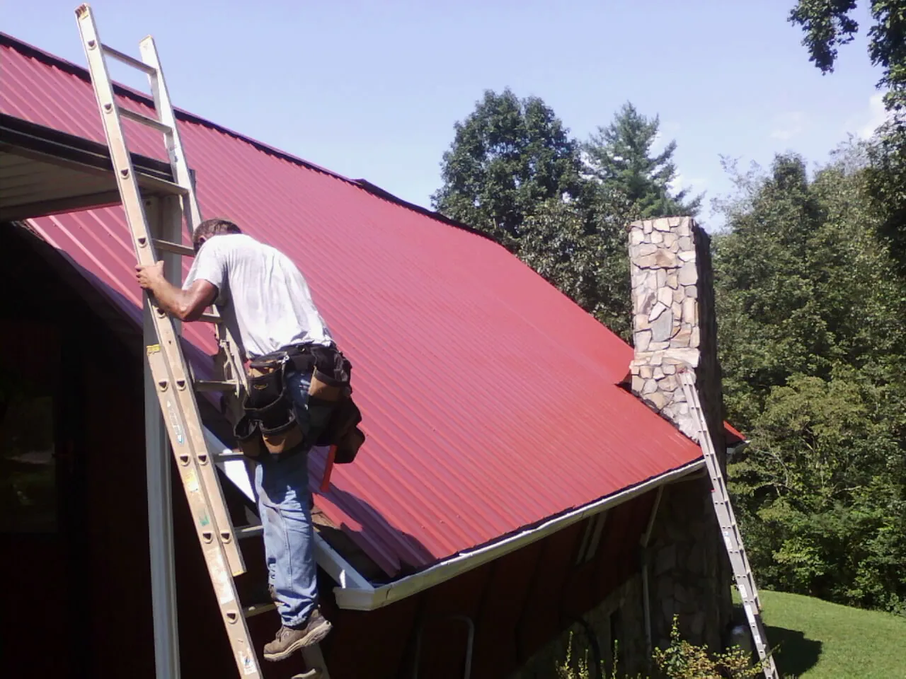 Roofer on a ladder beside a red metal roof and stone chimney — Soffit & Fascia Repair by Roofing Brothers in Alliance