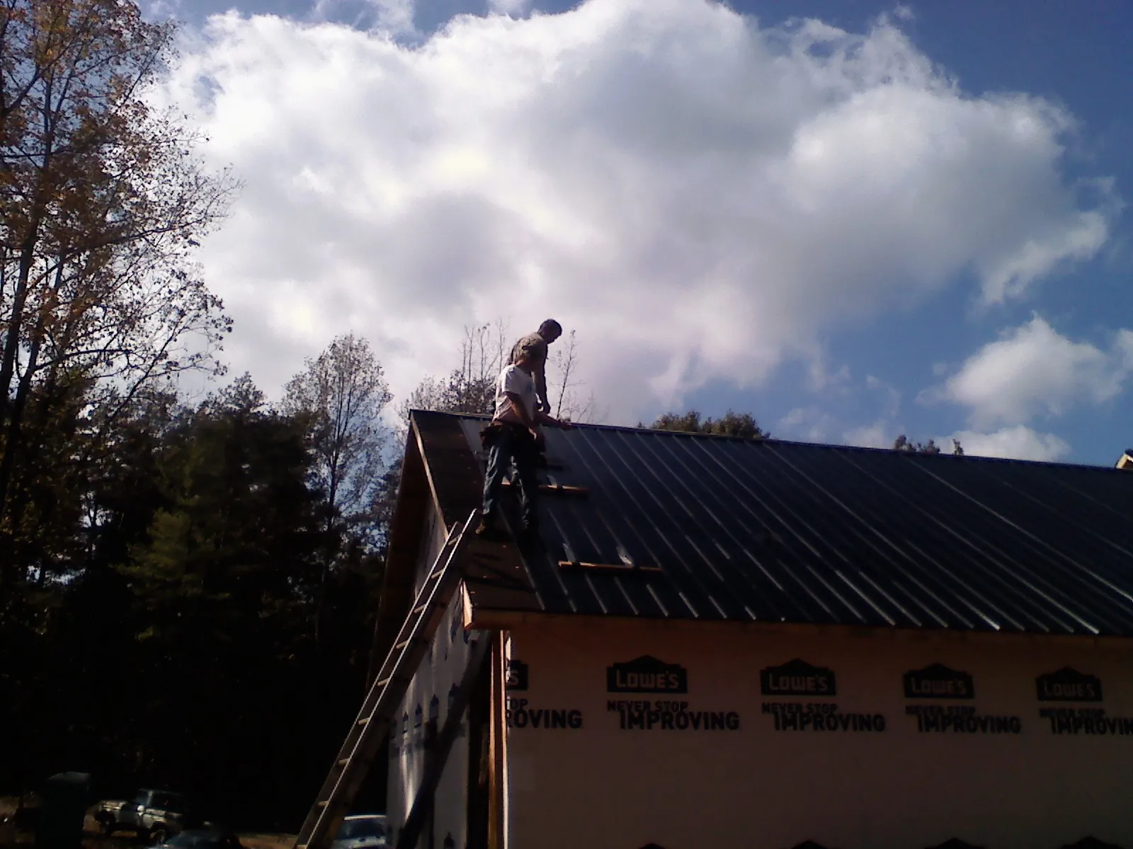 Roofer fastening dark metal panels high against a cloudy sky, from a Roof Inspection job in Alliance